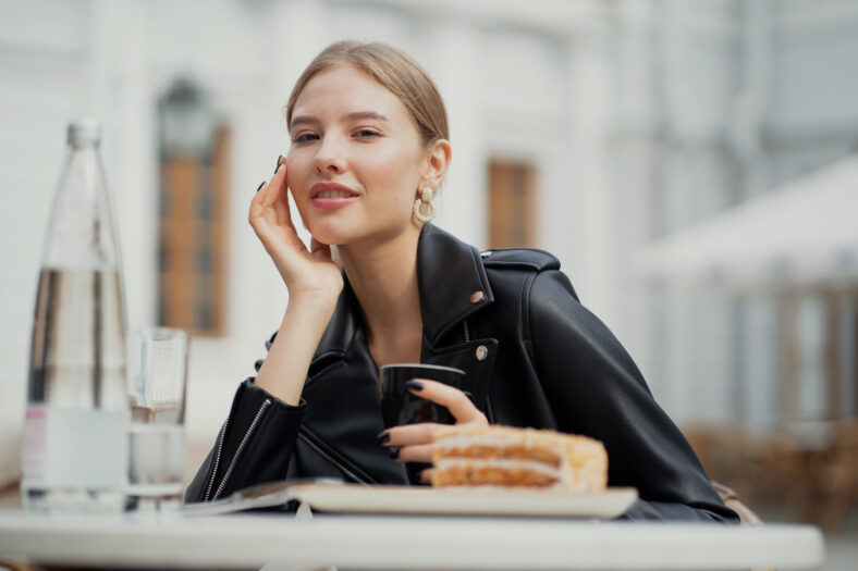 enjoying a delicious coffee, a young woman of European appearance ordered Breakfast, a Cup of Americano and delicious sugar desserts cake. black leather jacket, straight blonde hair.