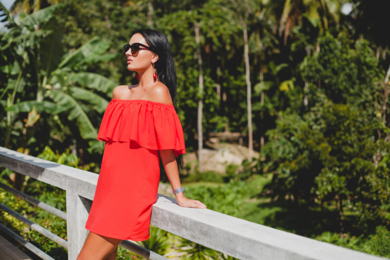 young stylish woman in red summer dress standing on terrace in tropical hotel, palm trees background, long black hair, sunglasses, ethnic earrings, sunglasses, smiling