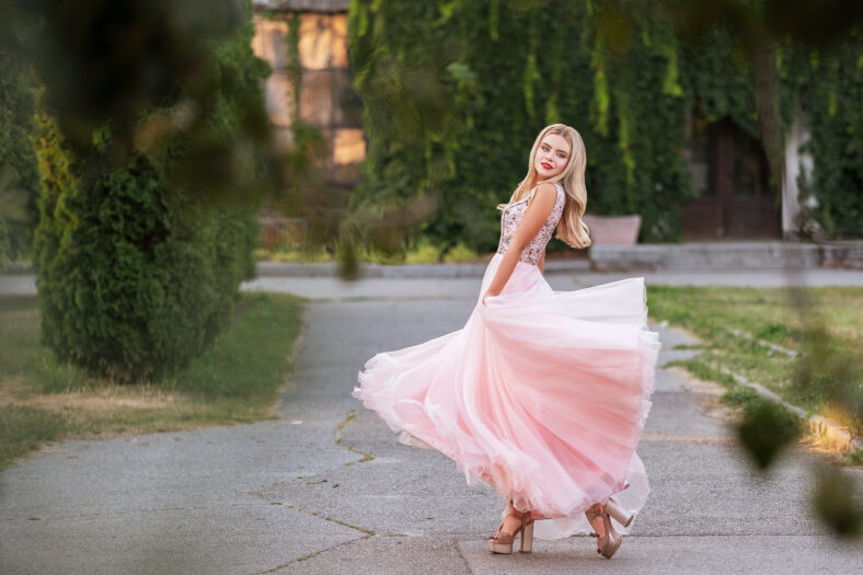 Blonde woman with make up in evening dress at park. The skirt of her dress fly on a wind. Sensual fashion portrait with a sunset light.