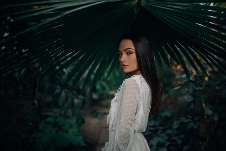 Portrait of young woman under big leaf of palm tree inside orangery.