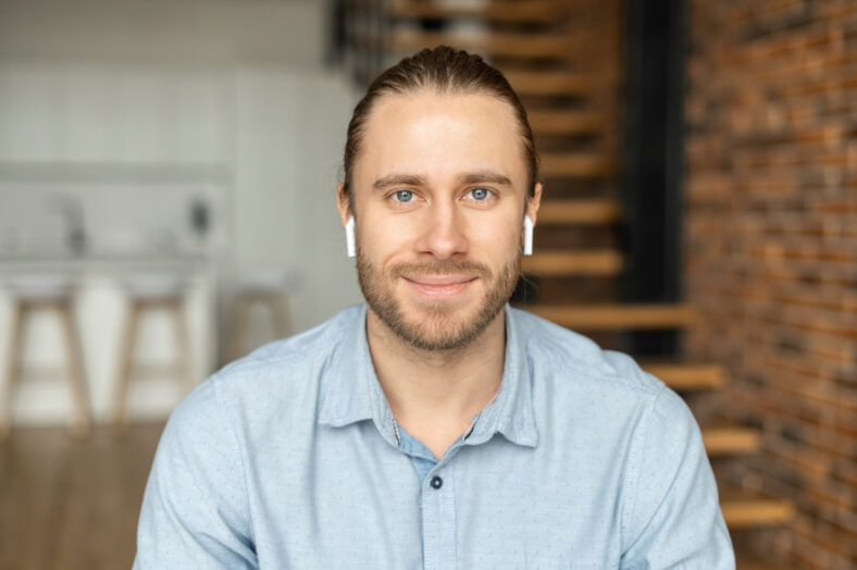 Portrait of an attractive young bearded hipster man smiling at the camera, wearing headphones, standing in the modern house, wearing a casual shirt. The view while the video-calling concept