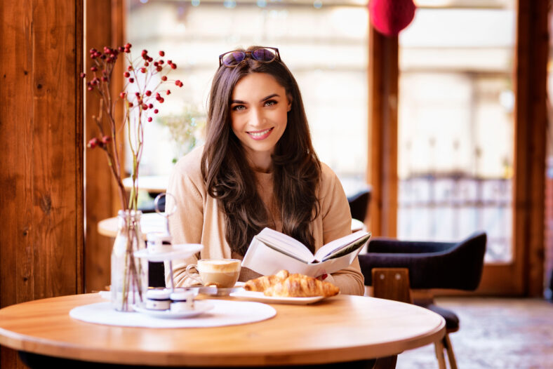 Portrait of beautiful young woman having breakfast and reading book while sitting at cafe desk.