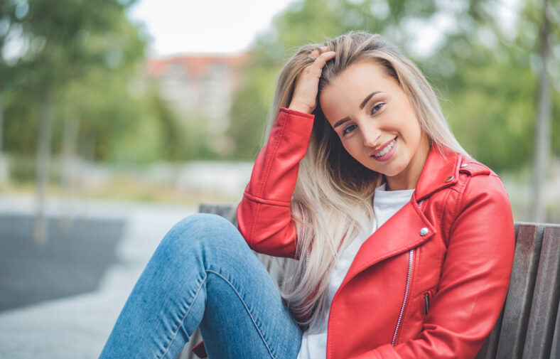Smile young beautiful woman sitting on bench. Outdoors daylight. Smiling pretty girl wearing casual clothing. Outside