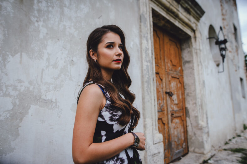 Beautiful brunette in dress goes in the courtyard of the old castle. Stylish girl in black dress on background of gray vintage building. Big sad eyes.