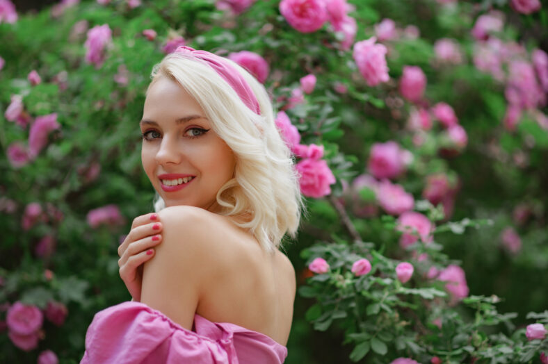 Happy smiling beautiful blonde woman wearing pink headband posing in blooming rose garden. Copy, empty space for text