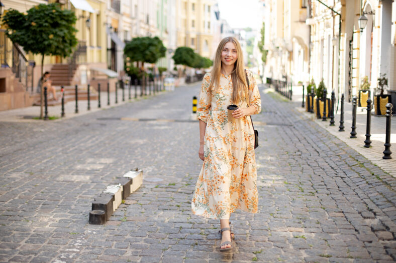 smiling caucasian woman blonde in dress with coffee takeaway walks on the cobblestones against the backdrop of the street and green trees in summer