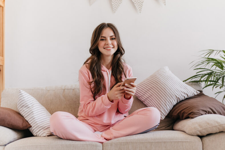 Photo made in homely atmosphere. Cute brunette in pink pajamas uses her phone and smiles sweetly