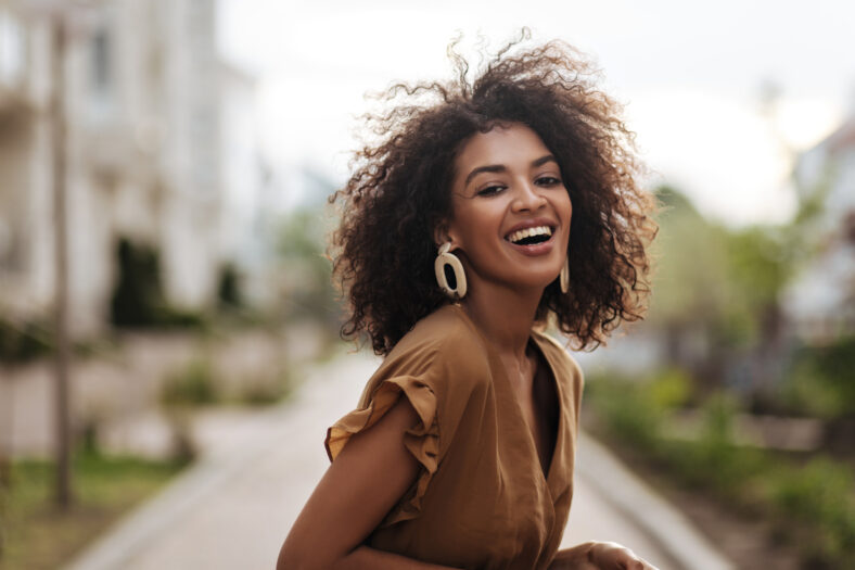 Cheerful brunette woman in brown dress smiles sincerely outdoors. Happy lady laughs and walks in great mood outside.