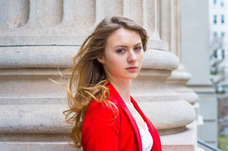 Vestirse con un blazer rojo y una camisa blanca, una adolescente rubia está de pie junto a una columna en un día ventoso, cuidadosamente mirándote.