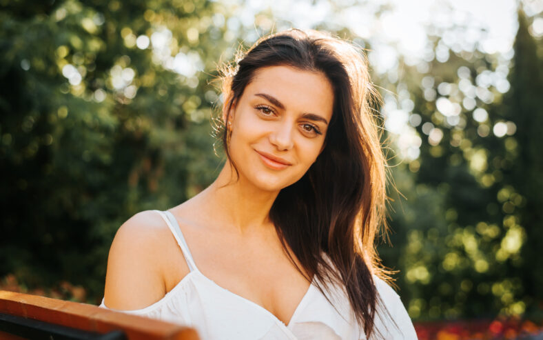 Portrait pretty young brunette woman in white light dress with gentle smile relaxing outdoors and looking at the camera. Beautiful Indian girl with brown big eyes summer lifestyle, close up.