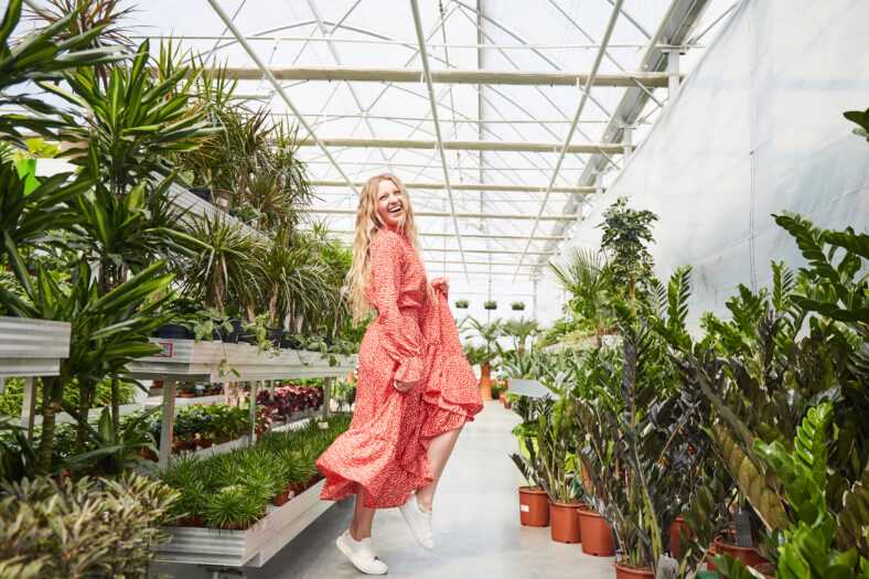 Young blond woman in red dress dancing into a greenhouse. Industrial Greenhouse with Flowers in pots all around.