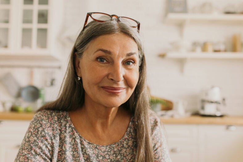Pretty grey-haired elderly woman keeping glasses on forehead posing in front of camera with adorable smile, wearing flowered dress, sitting against modern bright kitchen room. Lifestyle concept