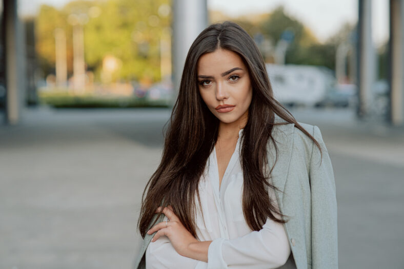 Portrait of beautiful attractive woman brunette with long hair dark eyes looking confidently into camera, arms crossed, wearing white shirt, elegant jacket over her back, blogger, businesswoman
