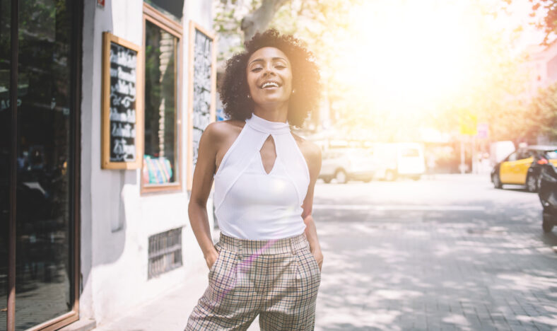 Cheerful young African American female in elegant clothes laughing while standing on street and looking at camera in summer sunny day