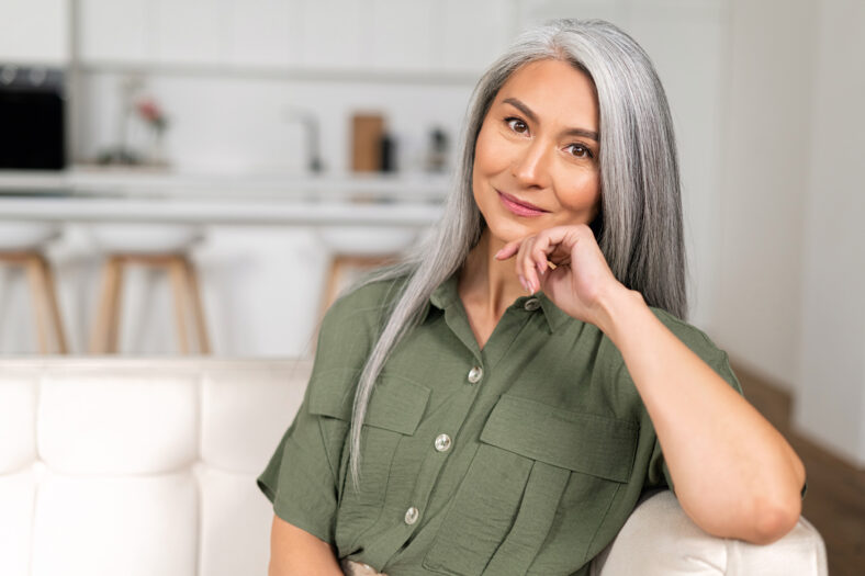 Close-up portrait of charming middle-aged woman with long silver hair sitting in relaxed athmosphere, mature 50s asian female resting at home, looking at camera and smiling with calm smile