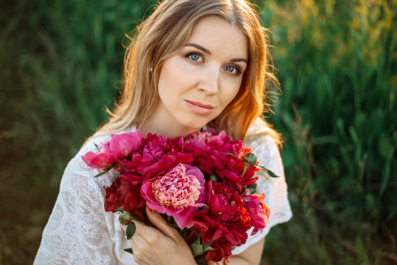 Portrait of an attractive young blonde woman in a white dress with a bouquet of pink peonies.