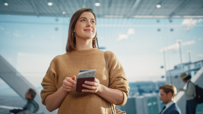 Airport Terminal: Happy Traveling Caucasian Woman Waiting at Flight Gates for Plane Boarding, Uses Mobile Smartphone, Checking Trip Destination on Internet. Smiling White Female on Vacation Trip