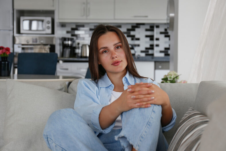 Portrait of young beautiful woman with long straight brunette hair sitting on the couch and smiling. Close up shot of brown eyed female with perfect smile. Copy space, background.