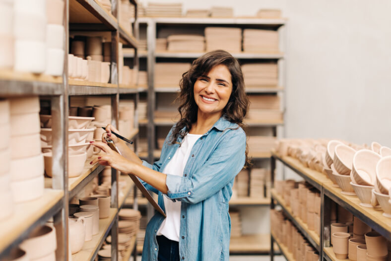 Cheerful ceramist smiling at the camera while stocktaking in her shop. Creative businesswoman managing a store with handmade ceramic products. Female entrepreneur running a successful small business.