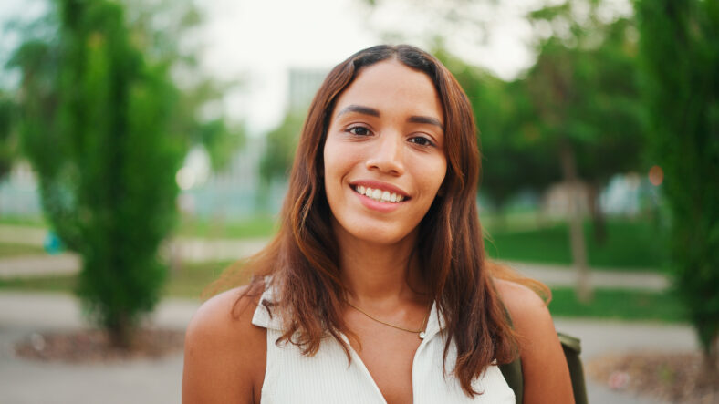 Clouse-up, beautiful girl with brown long hair, tanned skin wearing white top raises her head and looks straight into the camera.