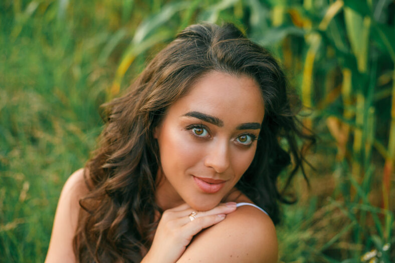 Delightful young woman in a white sundress in a field of green corn in the sunset light. Portrait of a beautiful model with dark long curly hair. Summer. Harvest. Eco. Bio.