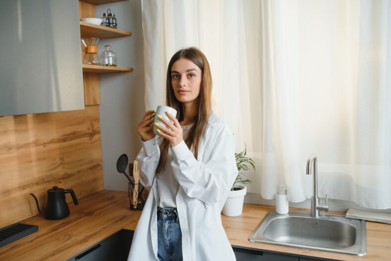 A young beautiful caucasian woman stands in the kitchen with a white cup of coffee or tea in the morning. A lonely girl is enjoying a cup of fresh hot drink