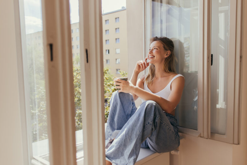 Cheerful young woman holding cup while sitting on windowsill at home,stock image
