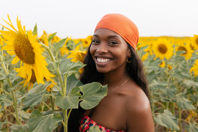 Feliz hembra negra en pañuelo naranja con cabello largo sonriendo y mirando a la cámara mientras está de pie cerca de girasol en el campo