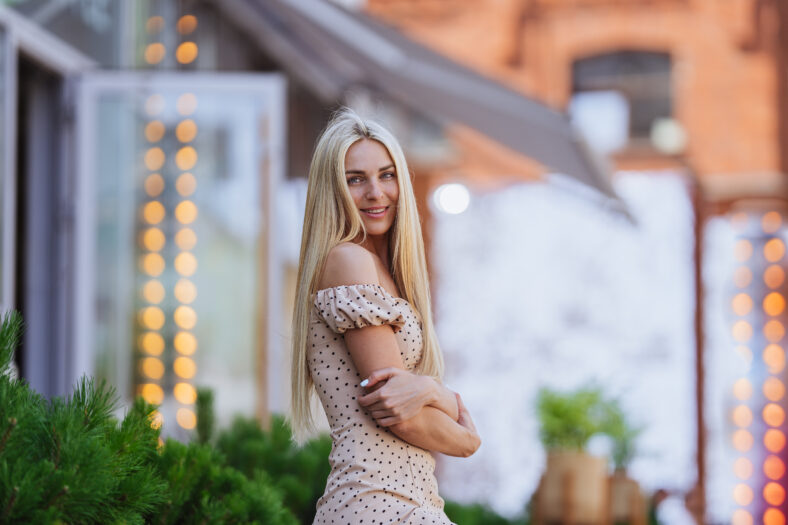 Gorgeous Blonde young woman on weekend waking outside looks at camera toothy smiling against blurry food court with plants. Stunning Swedish girl in love waiting for boy friend on Valentines day.
