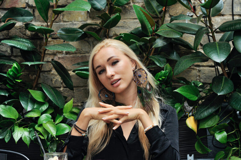 Close-up portrait of gorgeous blonde woman in boho chic dreamcatcher earrings jewelry with peacock feathers, Ficus elastica background loft apartment interior