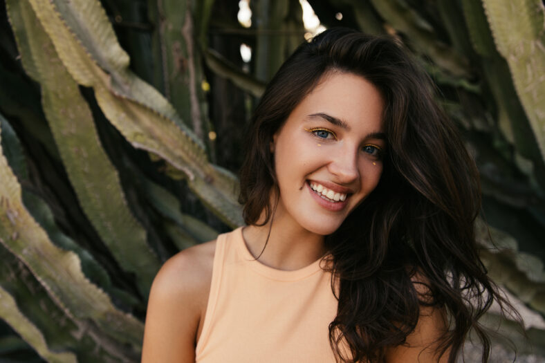 Close-up of positive young caucasian woman with stylish make-up looking at camera and smiles. Brunette wears peack color top. Concept of real positive emotions.