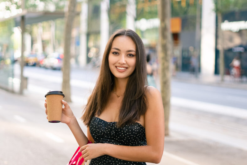 Happy young woman with paper bags and coffee to go smiling and looking at camera while standing on blurred background of city street after shopping in weekend