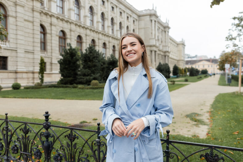 joyful young woman in blue trench coat standing near historical building in Vienna,stock image