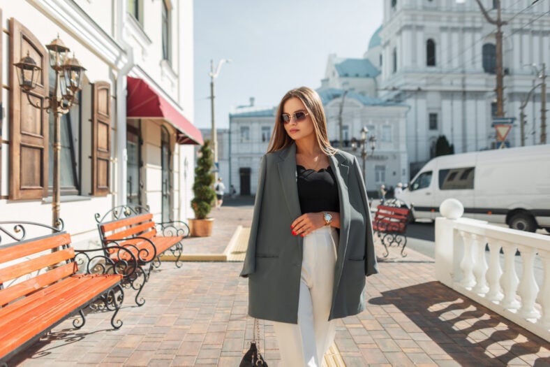 Stylish beautiful business lady with cool sunglasses in fashion casual clothes with a blazer walks in the city. Pretty chic urban woman in sunny spring day