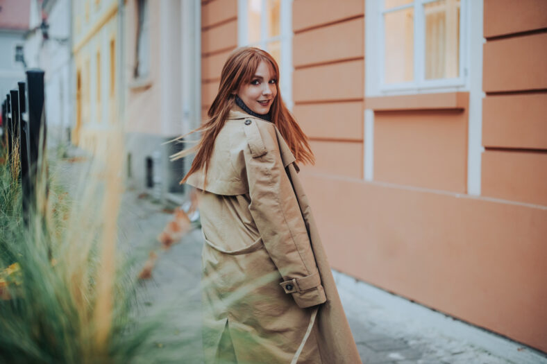 A fashionable ginger hair woman in vintage clothes is walking on city street downtown and looking over her shoulder at the camera. Portrait of chic young lady in retro clothes is smiling at the camera