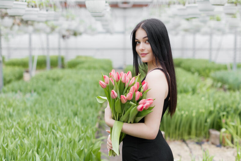 Young woman standing in large greenhouse and holding a blooming tulips in her hands.