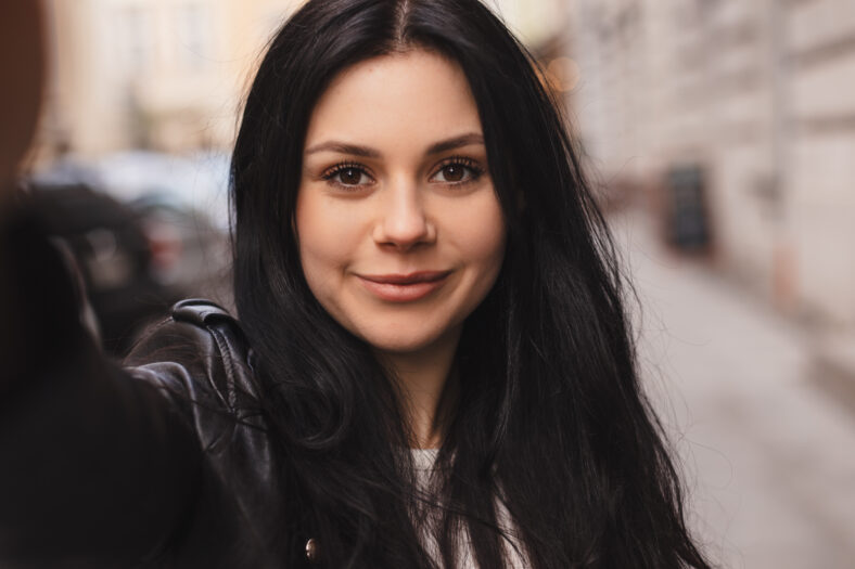 Close up portrait of young smiling woman tourist posing, walking in old town. Happy young model makes photo holding camera in hand close-up. Outdoor selfie photo in fashion outfit while walking.