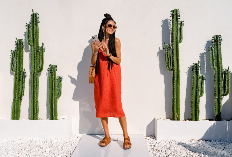 stylish smiling pretty woman with african braids on sunny summer vacation in colorful outfit wearing orange dress and sunglasses posing against white wall with cactuses in Mexico, using smartphone
