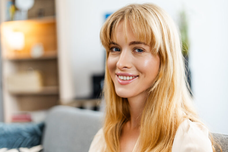 Portrait of a beautiful young woman with long blonde hair smiling while sitting on a comfortable sofa in her living room. Advertisement concept