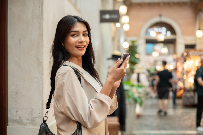An attractive, happy Asian woman in fashionable clothes stands in a shopping street in the city, holding her smartphone and smiling at the camera.