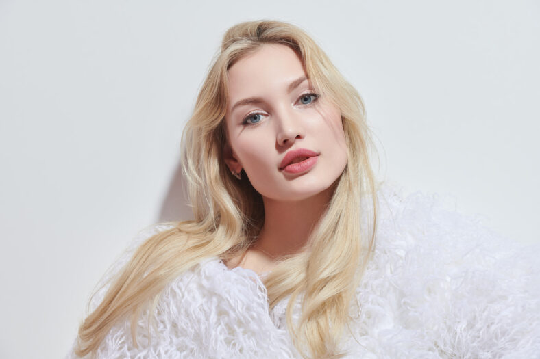 Fashion Shoot. Hermosa mujer joven con cabello rubio y labios posa en un esponjoso abrigo de piel blanca sobre un fondo de estudio blanco. Cosméticos de cuidado de belleza.