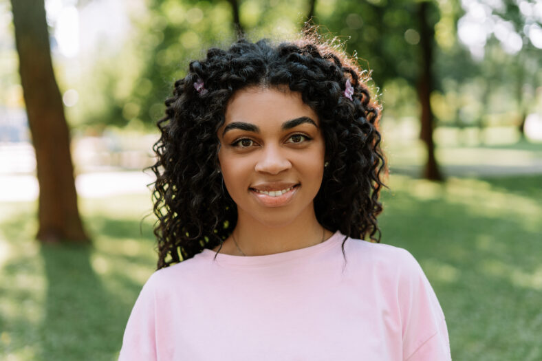 Portrait of african american smiling woman wearing stylish clothes looking at camera standing in green park. Positive lifestyle, natural beauty concept