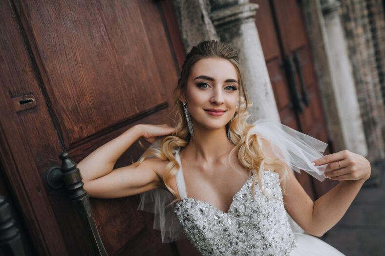 Beautiful young smiling bride curly blonde in white lace long dress stands, walks against the background of an old retro church, wooden doors. Wedding photography, portrait.