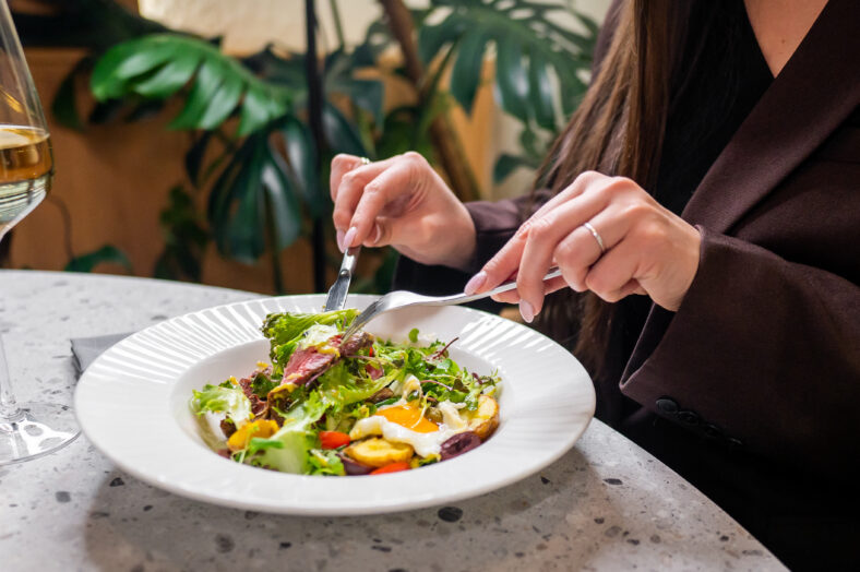 A person elegantly dining, using a fork and knife to enjoy a fresh salad. A glass of white wine is nearby, set in a stylish, modern indoor environment with greenery in the background.