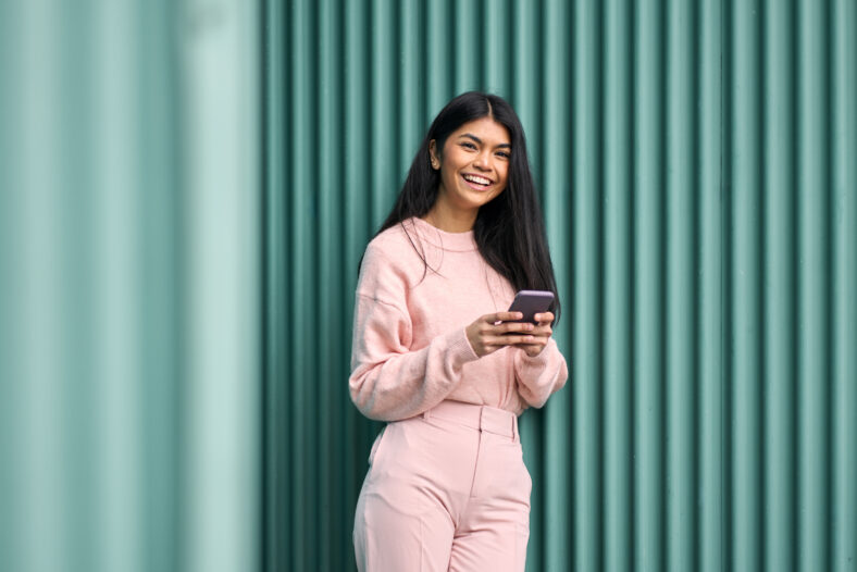 Feliz chica adolescente asiática con atuendo rosa usando aplicaciones de teléfonos móviles al aire libre mirando la cámara. Modelo femenino y sonriente positivo que sostiene el teléfono inteligente riendo de pie con fondo de pared azul.