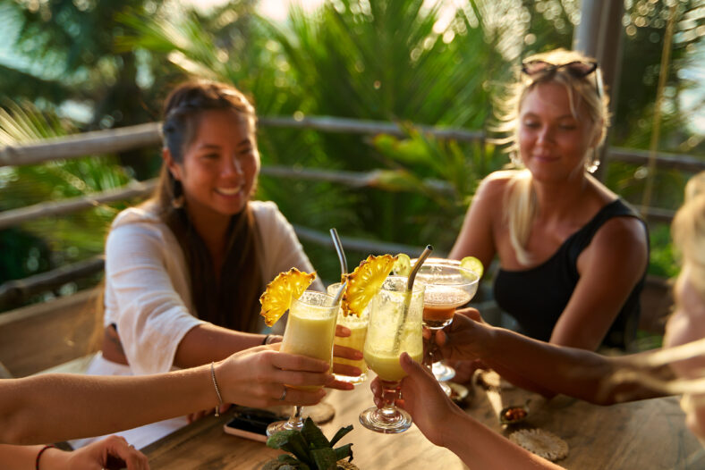 Diverse group of women at tropical restaurant toast with cocktails. Celebration. Vacation mood. Fun bachelorette party, on travel. Laughter in paradise. Enjoying sun vibes together. Happy moments.