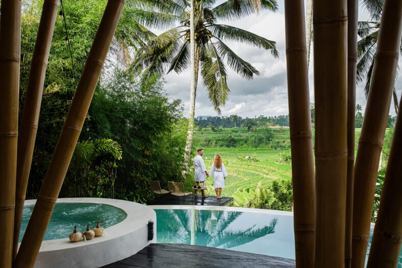 Romantic Couple in Bathrobes Enjoying Scenic Tropical Landscape from Luxury Infinity Pool Overlooking Rice Fields and Palm Trees in Bali, Indonesia.