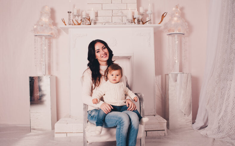 Smiling mother sitting with baby girl in armchair at home. Looking at camera. Motherhood.