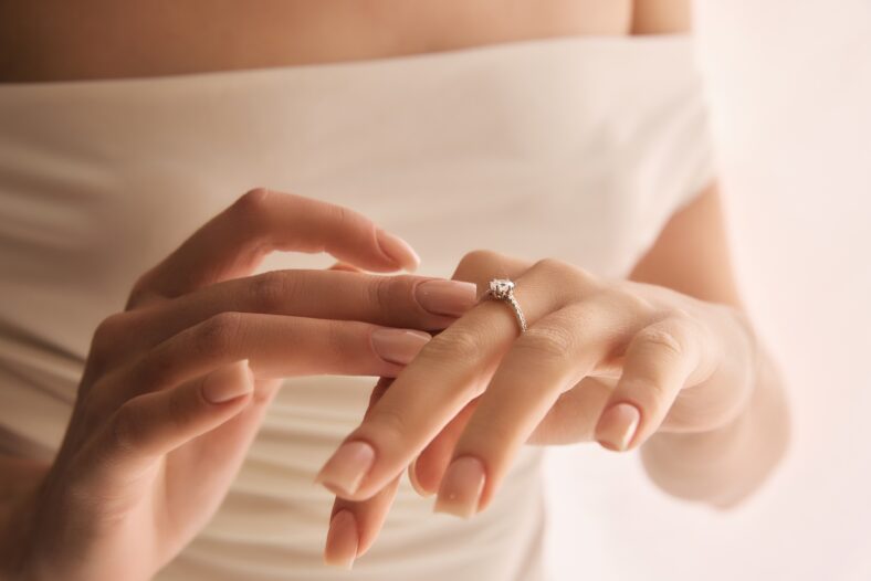 Close-up of female hands gently placing engagement ring with prominent stone onto ring finger against soft, light background. Concept of jewelry, luxury, accessories, wedding