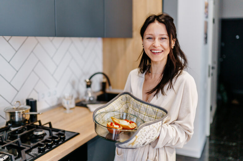 Mother in her 40s smiling and holding baked pears in glass dish, standing in modern home kitchen. Joyful moment after home cooking. Concept of healthy lifestyle, positive motherhood, simple pleasures.
